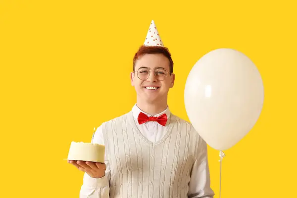 Young man in party hat with air balloon and sweet cake celebrating Birthday on yellow background