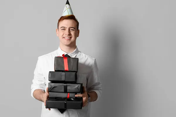 Happy young man in party hat with gift boxes celebrating Birthday on grey background