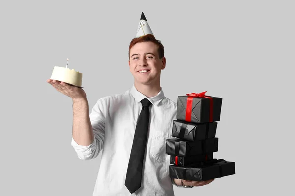 Happy young man in party hat with sweet cake and gift boxes celebrating Birthday on grey background