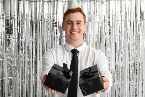 Young man in party hat with gift boxes celebrating Birthday on tinsel background