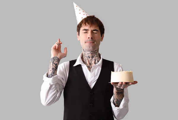 Young man in party hat with sweet cake celebrating Birthday on grey background
