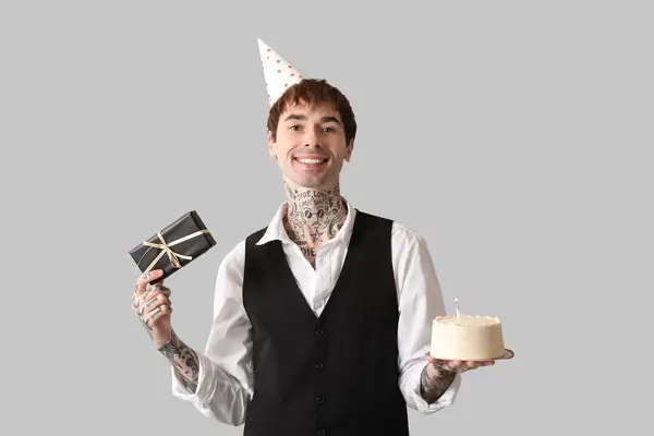 Young man in party hat with sweet cake and gift box celebrating Birthday on grey background