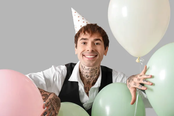 Happy young man in party hat with air balloons celebrating Birthday on grey background
