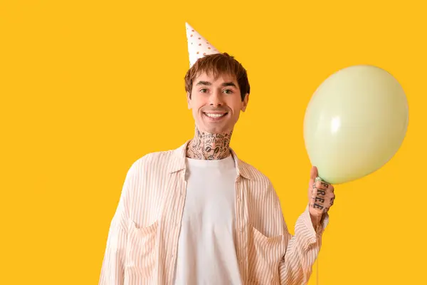 Young man in party hat with air balloon celebrating Birthday on yellow background
