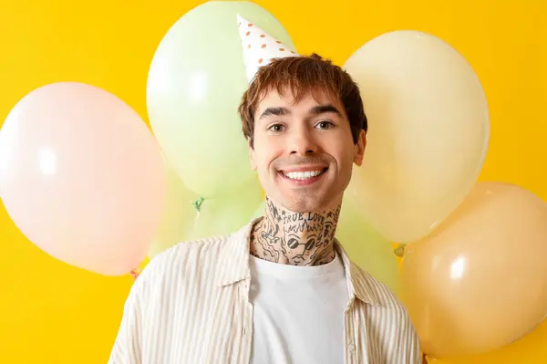 Young man in party hat with air balloons celebrating Birthday on yellow background
