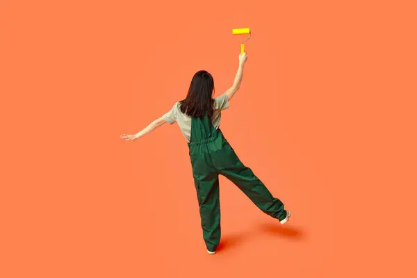 Female Asian painter with roller on orange background, back view