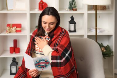 Evdeki koltukta otururken Noel tebrik kartı okuyan mutlu genç kadın.