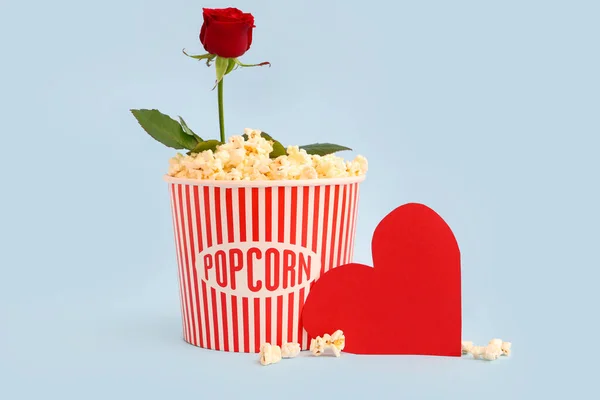Bucket of popcorn with beautiful rose and paper heart on blue background. Valentine\'s Day celebration