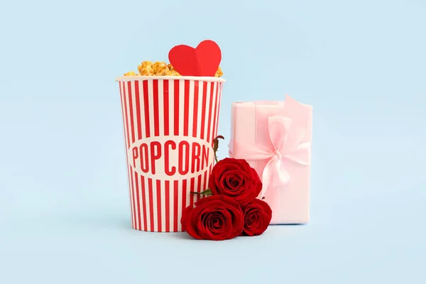 Bucket of popcorn with paper heart, gift box and beautiful roses on blue background. Valentine\'s Day celebration