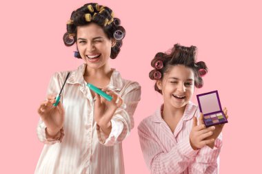 Küçük kız ve annesi pijamalarıyla pembe arka planda makyaj yapıyorlar.