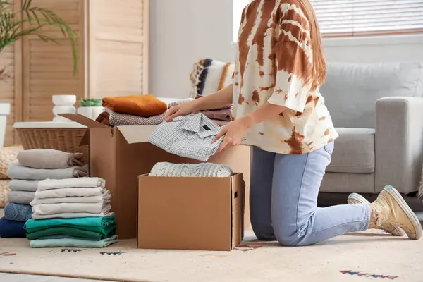 Young woman packing clothes into cardboard box in living room