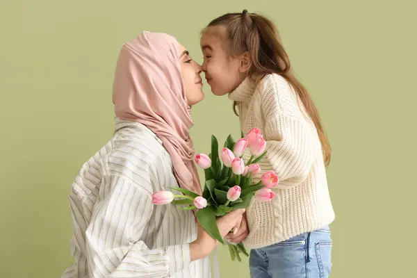 Young Muslim woman with tulips and her little daughter touching noses on green background. Mother\'s Day celebration