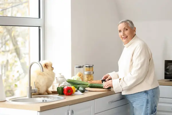 Senior woman with Pomeranian dog cutting vegetables in kitchen