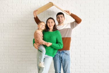 Çatı şeklinde mukavvalı olan mutlu bir aile beyaz tuğlalı arka plandaki yeni evlerini düşlüyor.