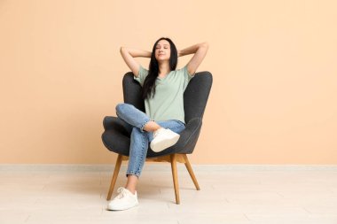 Güzel genç bayan bej duvarın yanındaki rahat koltukta dinleniyor.