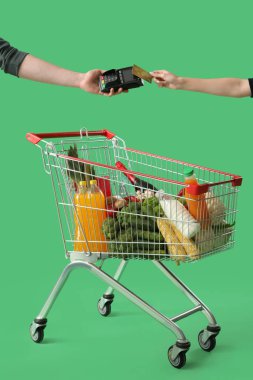 Yeşil arka planda yiyecek dolu alışveriş arabasının parasını ödeyen kadın.