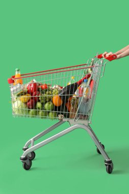 Yeşil arka planda farklı yiyeceklerle dolu alışveriş arabasını iten kadın.