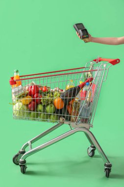 Yeşil arka planda yiyecek dolu alışveriş arabasının yanında ödeme terminali olan bir kadın.