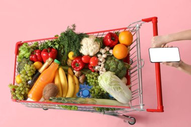 Cep telefonlu bir kadın pembe arka planda yiyecek dolu alışveriş arabalarını itiyor.