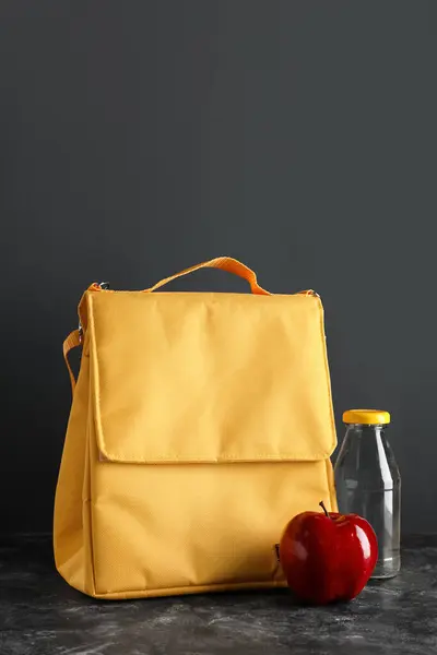 Lunch box bag with apple and bottle of water on grunge table near black wall