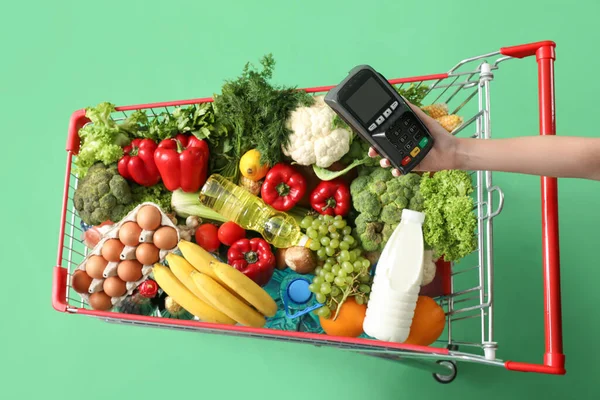 Ödeme terminali olan bir kadın yeşil arka planda yiyecek dolu alışveriş arabalarını itiyor.