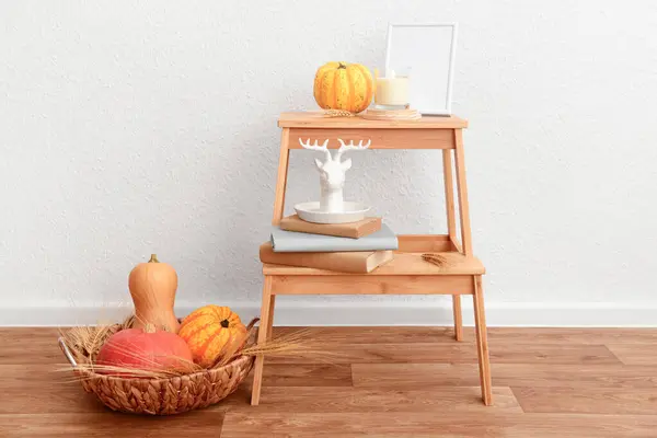 Wooden stepladder with pumpkins, books and candle near white wall