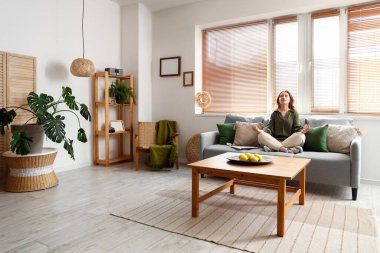 Modern oturma odasındaki gri kanepede meditasyon yapan genç ve güzel bir kadın.