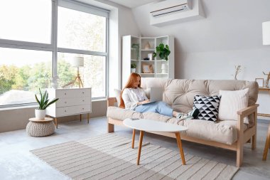 Genç ve güzel bir kadın, beyaz koltukta otururken dizüstü bilgisayar kullanıyor.