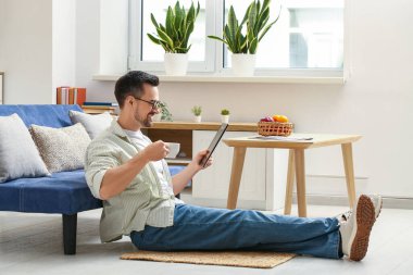 Elinde kahve fincanı olan yakışıklı adam oturma odasında tablet bilgisayar kullanıyor.