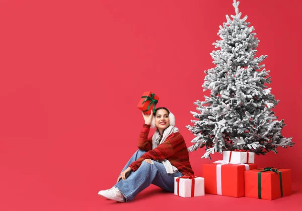 赤い背景にクリスマスツリーの近くにギフトボックスを持つ美しい若い女性 — ストック写真