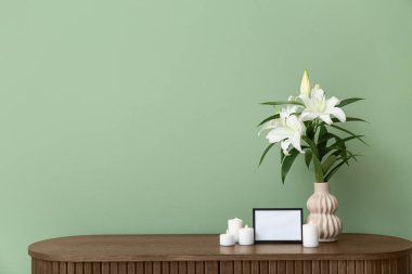 Boş cenaze çerçevesi, yanan mumlar ve vazo, ahşap dolapta, renkli duvarın yanında zambak çiçekleri.