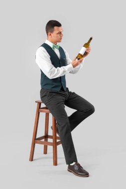 Elinde şarap şişesi olan genç barmen gri arka planda taburede oturuyor.