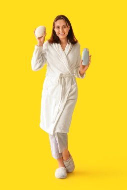 Bornozlu genç kadın duş jeli ve banyo lifiyle sarı arka planda.