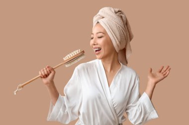 Bornozlu genç Asyalı kadın masaj fırçası ile kahverengi arka planda şarkı söylüyor.