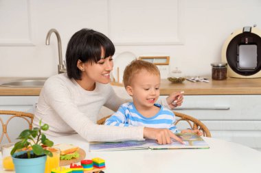 Küçük oğlu mutfaktaki masada kitap okurken mutlu bir anne.