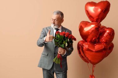 Elinde gül buketi ve kalp şeklinde hava balonları olan olgun bir adam bej arka planda baş parmağını gösteriyor. Sevgililer Günü kutlaması