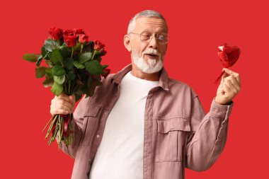 Kırmızı arka plandaki Sevgililer Günü için elinde gül buketi ve kalp şeklinde hava balonu olan olgun bir adam.