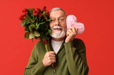 Kırmızı arka planda Sevgililer Günü için gül buketi ve kumaştan kalbi olan olgun bir adam.