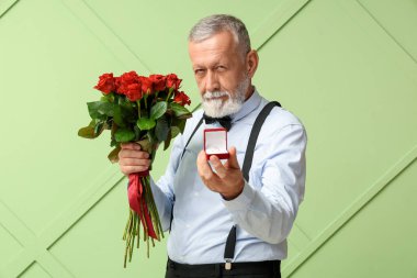 Elinde gül buketi ve yeşil arka planda nişan yüzüğü olan bir kutu tutan olgun bir adam. Sevgililer Günü kutlaması