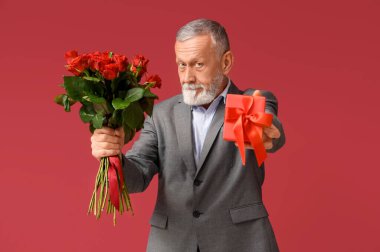 Kırmızı arka planda Sevgililer Günü için bir buket gül ve hediye taşıyan olgun bir adam.