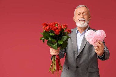 Kırmızı arka planda Sevgililer Günü için gül buketi ve kumaştan kalbi olan olgun bir adam.