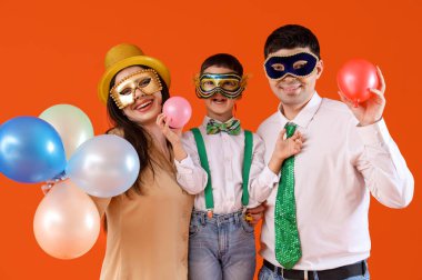 Kostümlü mutlu aile ve turuncu arka planda balonları olan karnaval maskeleri.