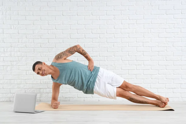 Young man with laptop doing yoga on mat near white brick wall