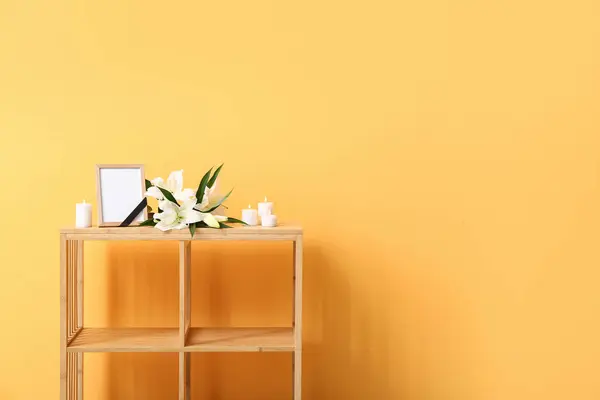 Blank funeral frame, burning candles and lily flowers on wooden shelf unit near color wall