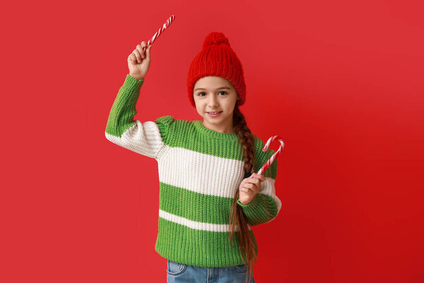 Cute little girl in warm hat with candy canes on red background