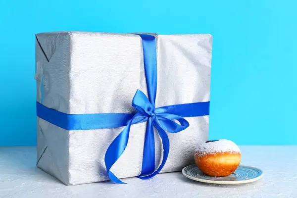 Tasty donut and gift box for Hanukkah celebration on table against color background