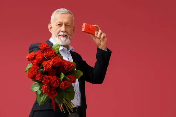 Mature man with bouquet of roses and gift for Valentine's day on red background