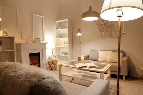 Interior of elegant living room with grey sofas, fireplace, white furniture and glowing lamps at evening
