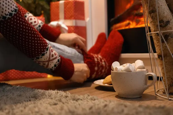Woman with warm socks and cup of hot chocolate sitting near fireplace in festive living room at evening