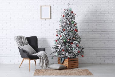 Beyaz tuğla duvara karşı hediyeler ve koltukla süslenmiş Noel ağacı.